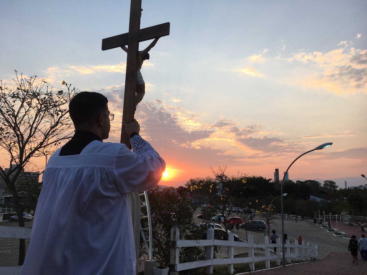 otografia em ângulo baixo de um jovem acólito, de costas, vestindo uma sobrepeliz branca sobre batina preta. Ele segura uma cruz de madeira com a imagem de Cristo crucificado, elevando-a em direção ao céu durante o pôr do sol. O sol está posicionado logo abaixo do braço da cruz, criando um brilho intenso. O cenário é um ambiente externo elevado, com uma cerca branca em primeiro plano, árvores com flores amarelas e uma vista de um estacionamento e prédios ao fundo sob um céu com tons de rosa e laranja.