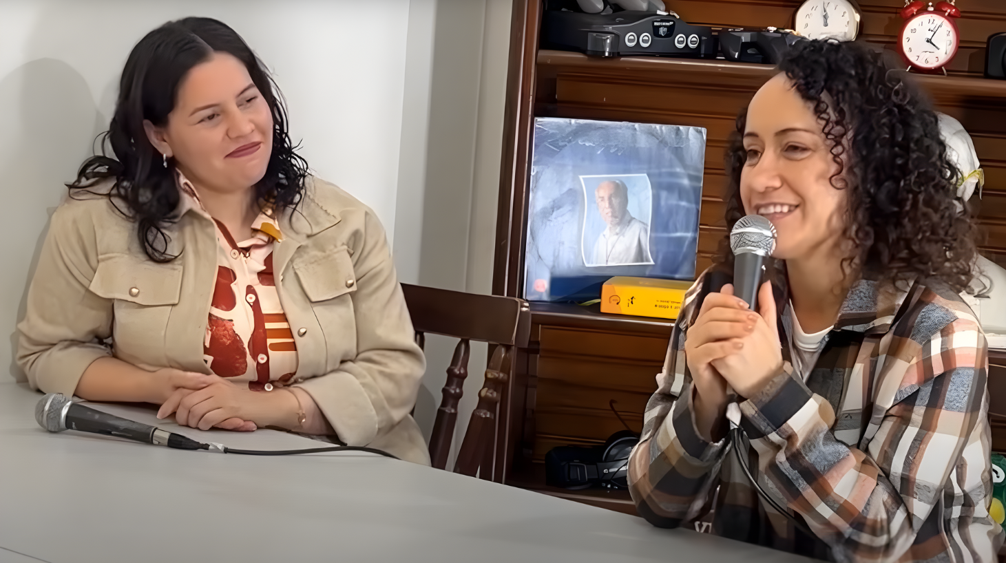 Ana Lúcia (à esquerda, com camisa estampada) e Maria Lídia/Marley (à direita, de camisa xadrez, segurando um microfone) sorrindo durante o podcast. Ao fundo, uma foto de Monsenhor Jonas Abib, reforçando o tema desafio de amor e vocação.