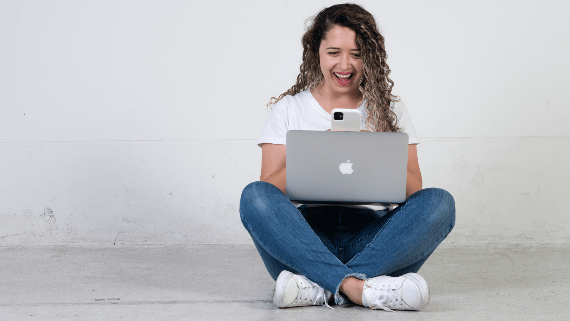 Jovem mulher entusiasmada com o projeto construindo, sentada no chão com notebook e celular em um fundo branco. Jovem missionária com cabelos cacheados, vestindo camiseta branca, calça jeans e tênis brancos, sentada no chão de pernas cruzadas em frente a uma parede branca lisa. Ela está olhando para um notebook prateado e segurando um celular branco com a outra mão. Sua expressão é de alegria e entusiasmo, ao fazer cadastro para o projeto construindo com a boca aberta em um sorriso.