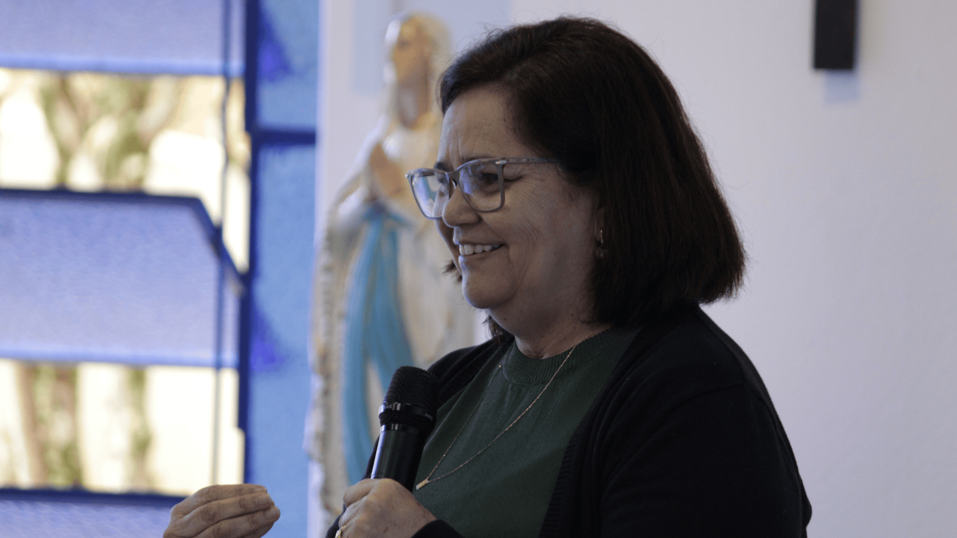 Uma foto de plano médio de Vera Lúcia Reis (uma mulher de pele clara, usando óculos de aro, blusa verde escura e casaco preto), sorrindo e falando em um microfone. Ela gesticula com a mão esquerda enquanto segura o microfone com a direita. Ao fundo, ligeiramente desfocada e atrás dela, é visível uma imagem da Virgem Maria, com o título de Nossa Senhora de Lourdes, vestida em um manto azul e branco. A cena acontece na Sala do Chamado na Canção Nova Queluz/SP em um ambiente interno com iluminação clara, em uma sala de reuniões.